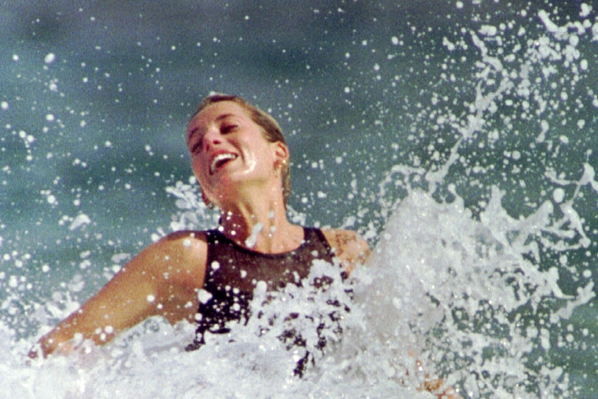 Princess Diana in a black swimsuit looking joyful as a wave breaks around her