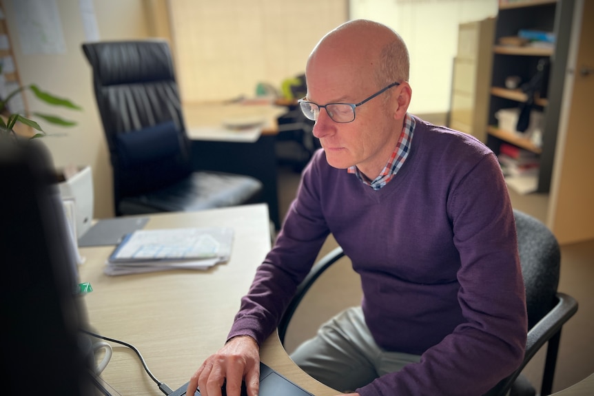 Tim sits in a chair at his desk, typing. He is wearing a purple jumper, grey pants and black glasses.