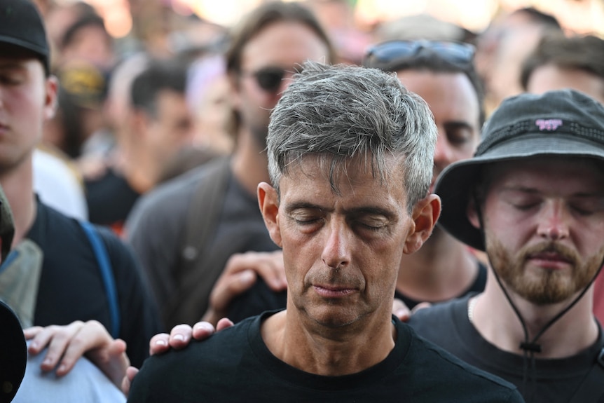 A man wearing a black t-shirt and a hat, places his hand on another man in a black t-shirt.
