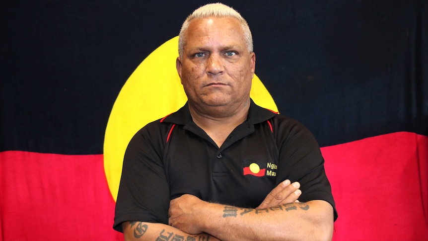 A man in a black polo shirt stands with arms crossed in front of an Aboriginal flag.