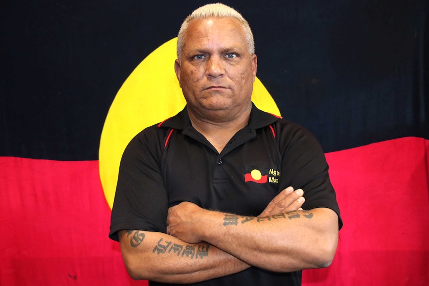 A man in a black polo shirt stands with arms crossed in front of an Aboriginal flag.