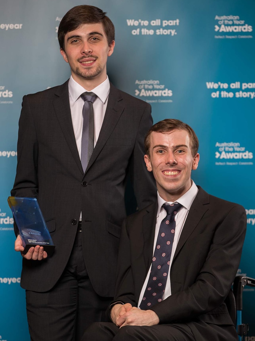 Two brothers wearing suits and one sitting in a wheelchair smile at the camera while holding a trophy in front of a banner.