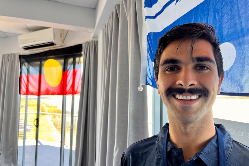 A young man with a moustache smiles at the camera with the Ngarrinderji and Aboriginal flags in the background