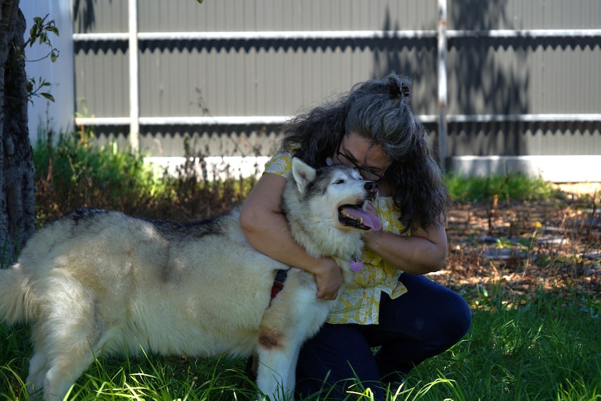 A woman with long grey hair with her arms around a senior husky dog in a backyard
