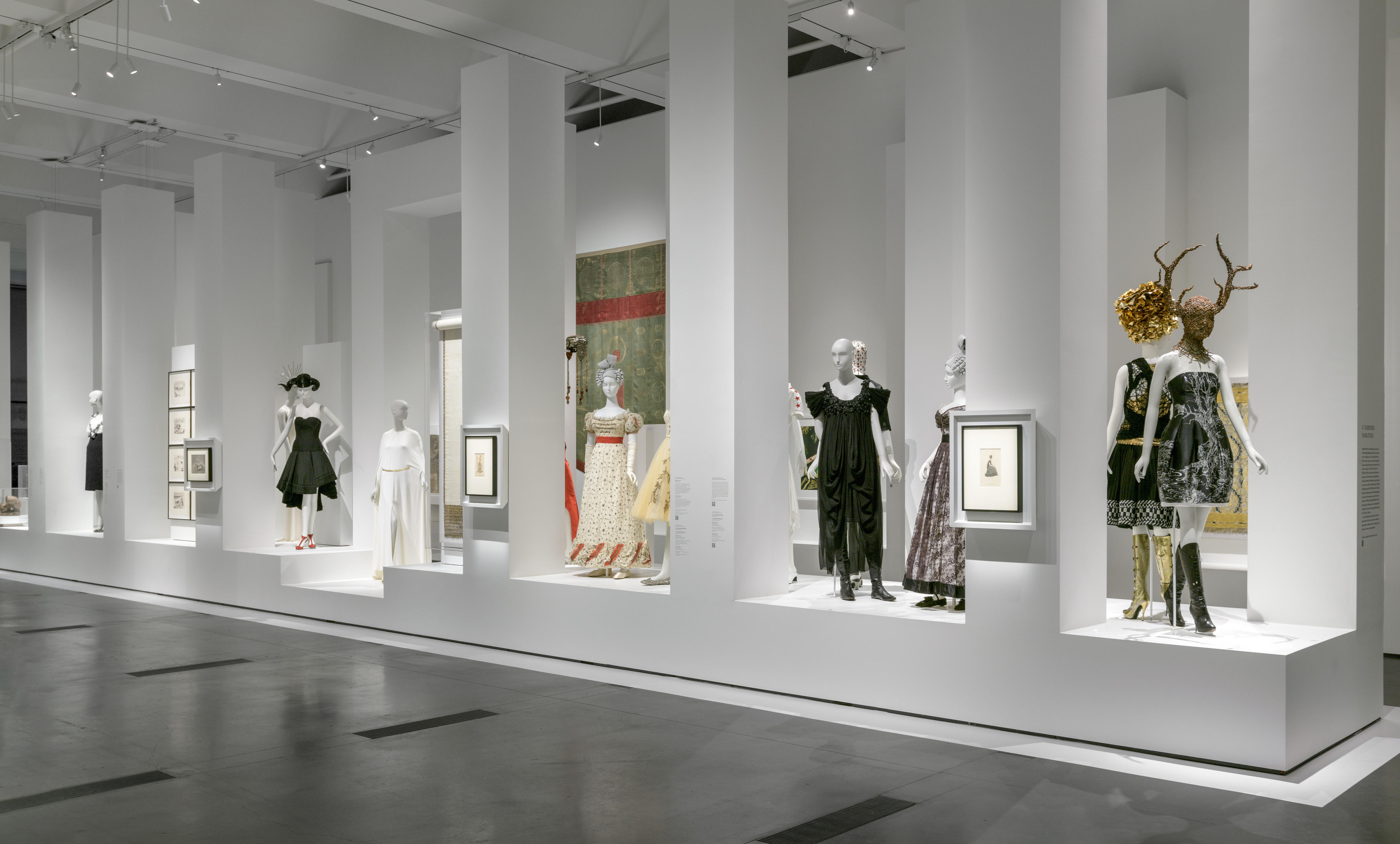 In einem Galerieraum stehen mehrere Schaufensterpuppen, die extravagante Kleider, Stiefel und Kopfbedeckungen tragen