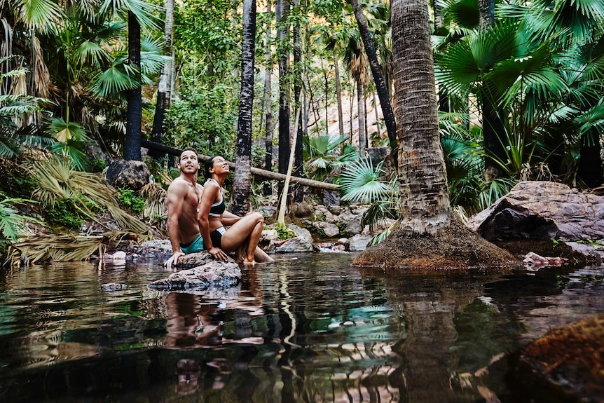 Two people sit in a spring surrounded by lush green plants.