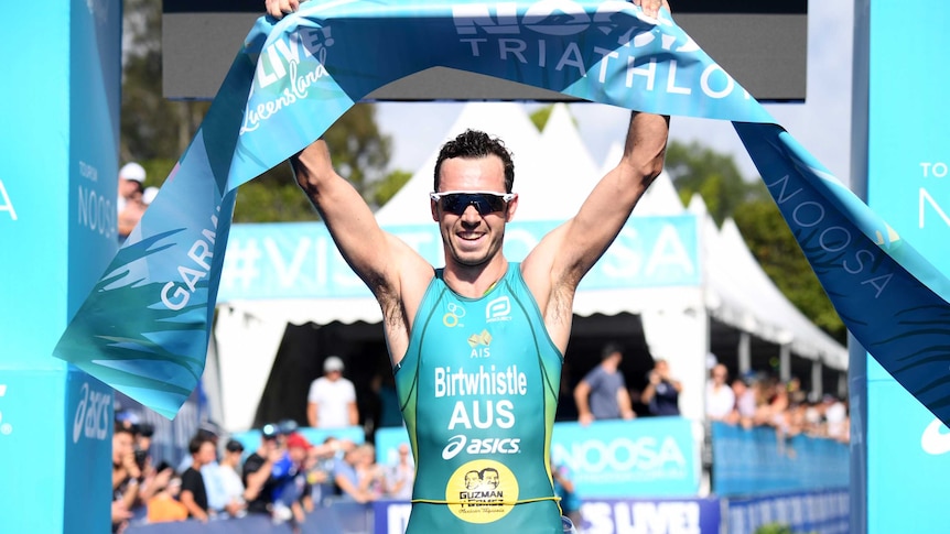 An athlete, wearing sunglasses and a skintight suit, raises his arms in victory as he crosses the line.