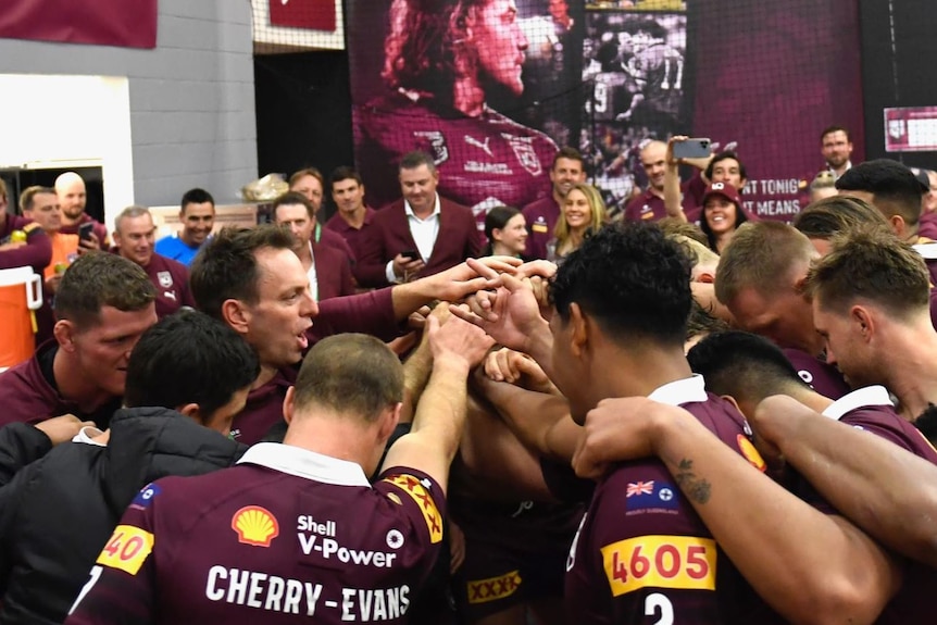 Maroons players in uniform in the locker room put their hands together in a close group circle 