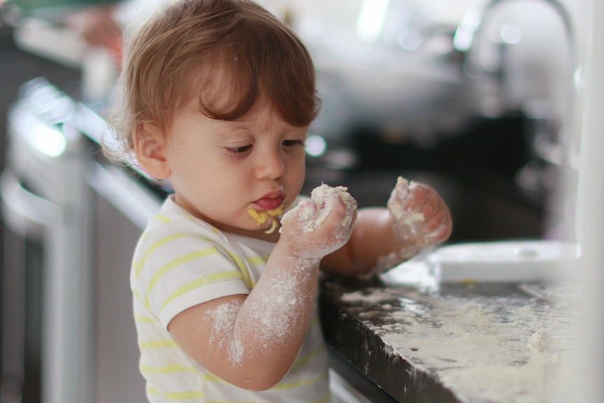 A baby plays with dough and flour at a kitchen bench, in an article about the importance of letting kids play with food.