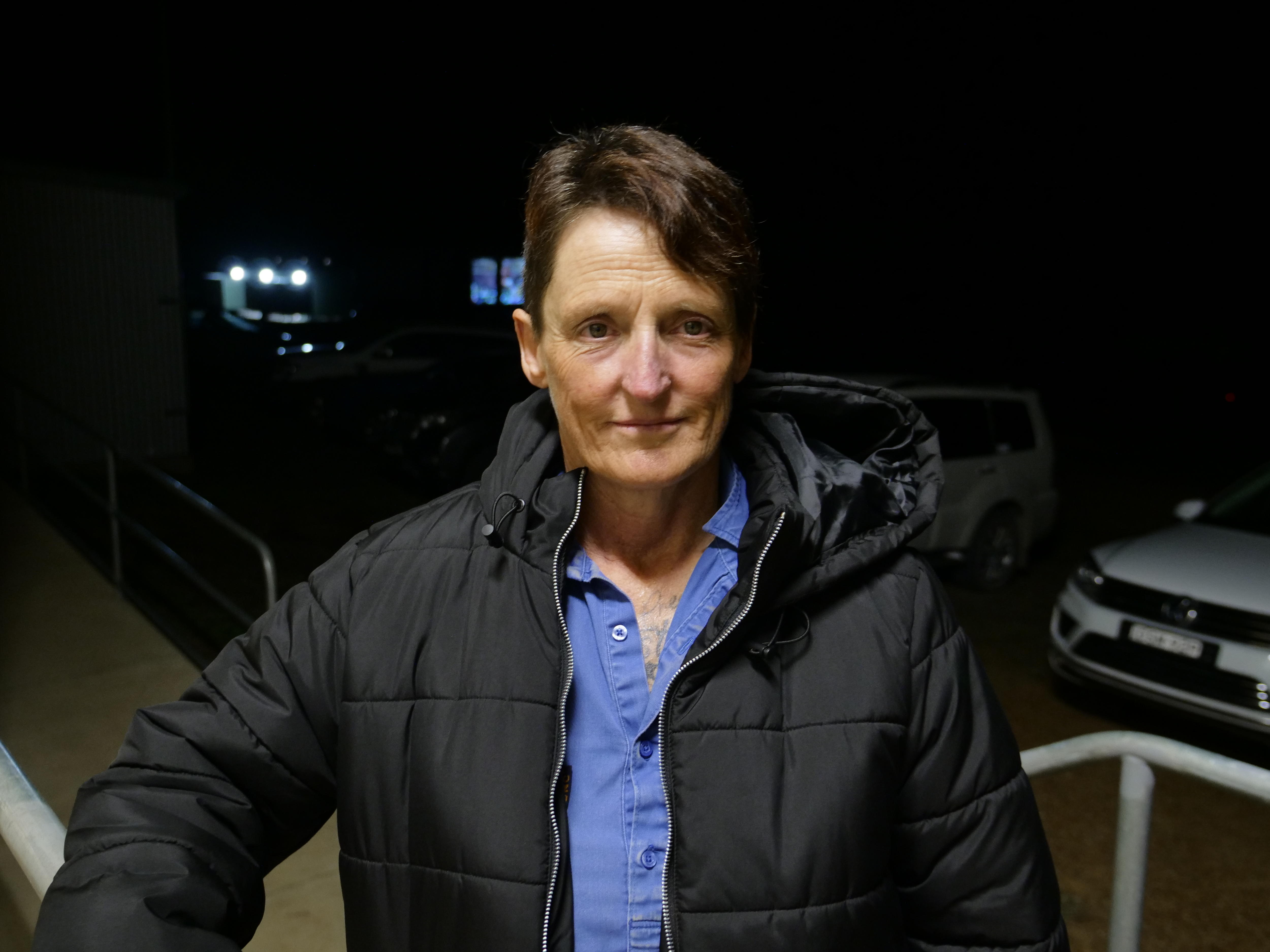 Sue with short hair gazes directly at the camera, background is dark, cars parked.