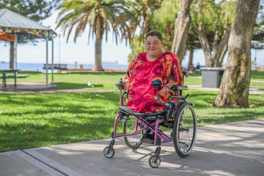 A woman with short hair, wearing red in a wheelchair in a park near a beach, who has had her legs and hands amputated.