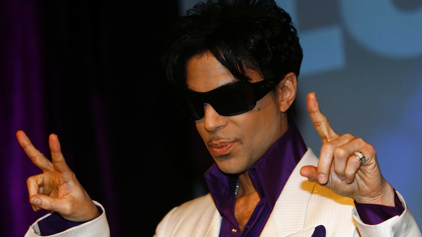 Prince wears sunglasses, a white suit and purple show while holding a peace sign