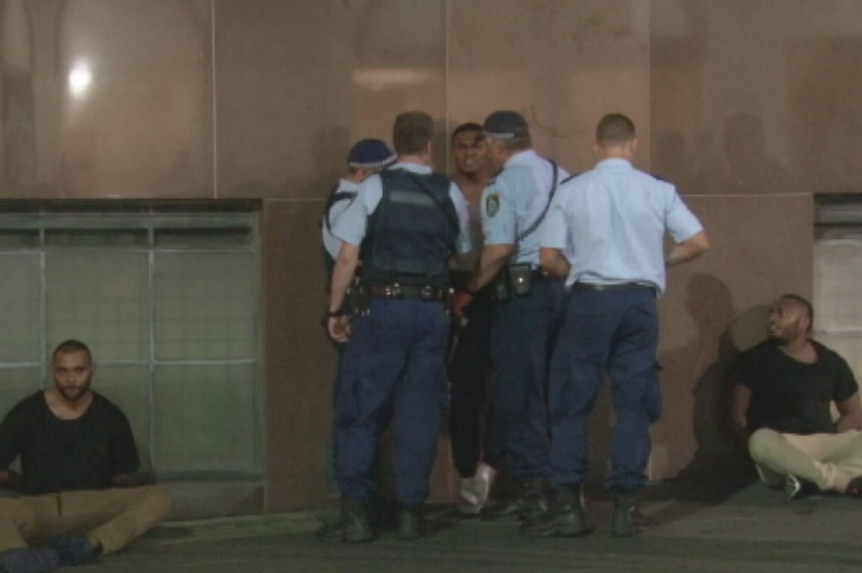 Two men sit on the ground with their hands behind their backs while a third man stands while being detained by police.