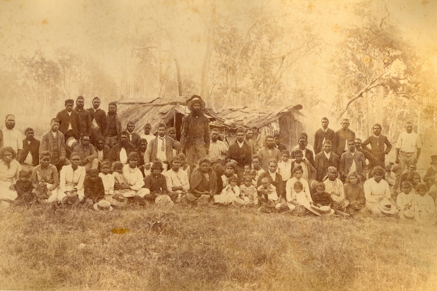 A sepia photo from the late 1800s of a group of Indigenous people dressed in clothes of that period stand in front of bark huts