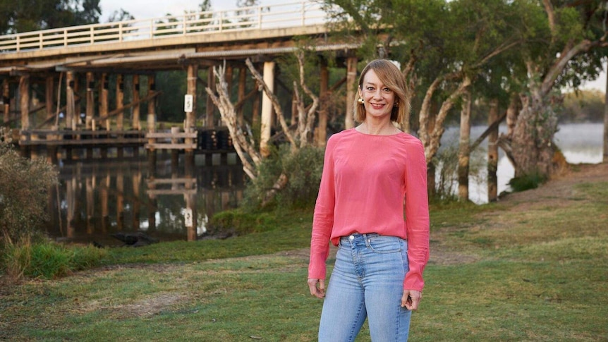 A woman with shoulder-length dark blonde hair, wearing a pink top and jeans, standing near a bridge over a river.