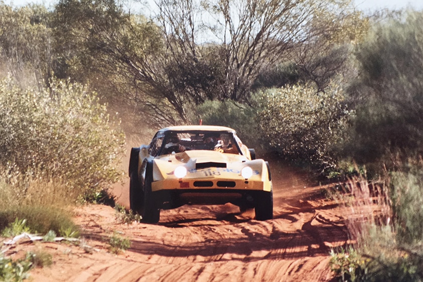 A modified car is driven through dirt and sand in outback terrain.