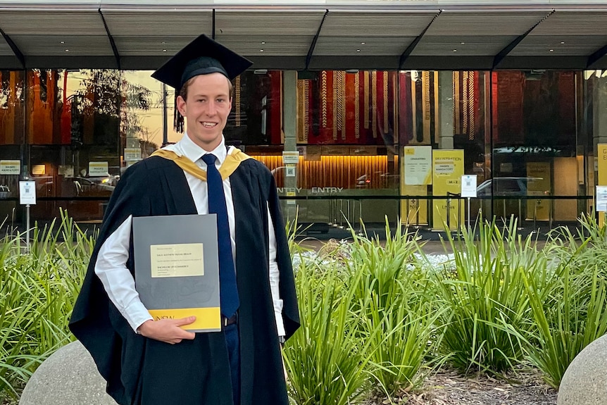 A man wearing academic regalia with a white shirt and blue tie, holds up his degree in front of UNSW. 