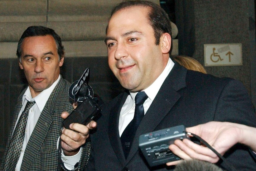Tony Mokbel, wearing a suit, smiles as reporters hold microphones in front of his face.