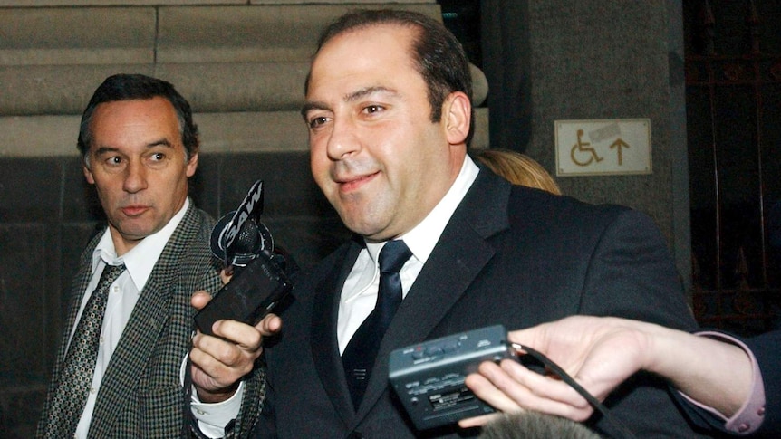 Tony Mokbel, wearing a suit, smiles as reporters hold microphones in front of his face.