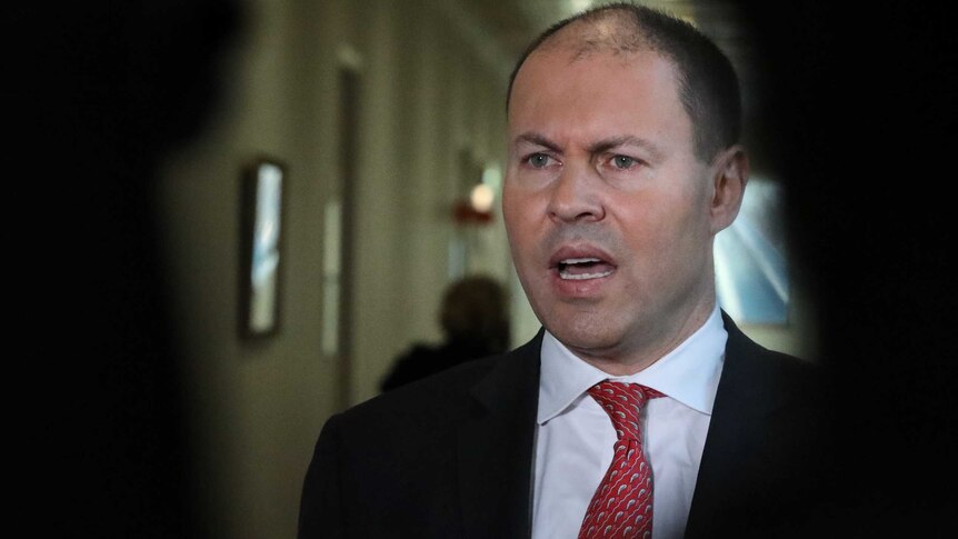 Josh Frydenberg wears an angry facial expression. He is surrounded by shadow.