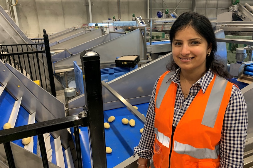 Indian/Australian lady with dark hair and an orange high vis vest stands in front of a potato conveyer-belt