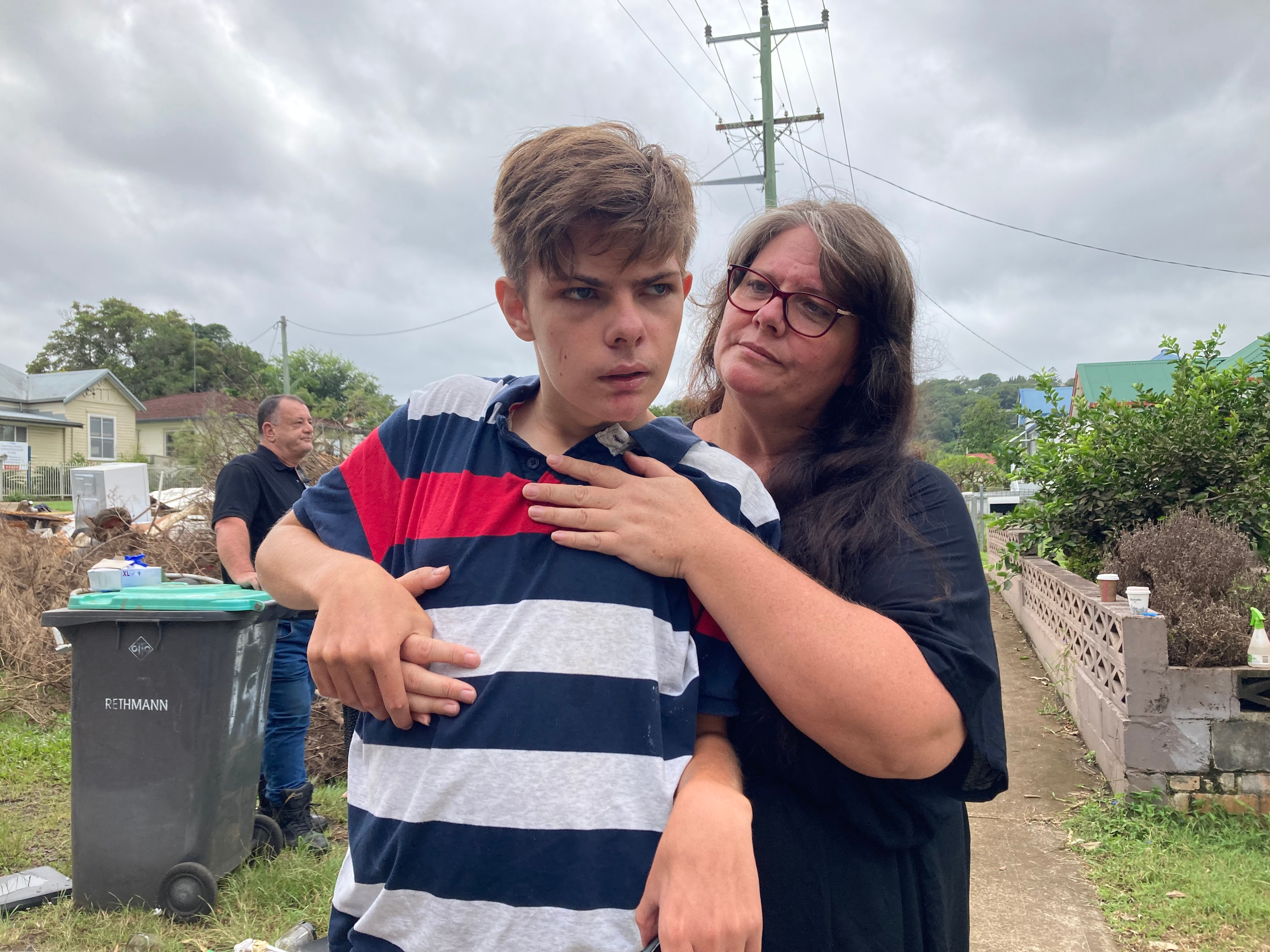 A woman hugs a teenage boy, standing in a suburban street.