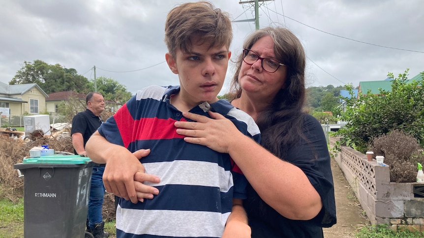 A woman hugs a teenage boy, standing in a suburban street.