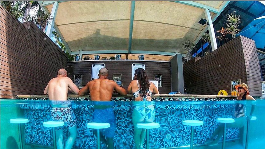 A semi-immersed image of four people in swimming costumes sitting on underwater barstools at a swim-up pool bar.
