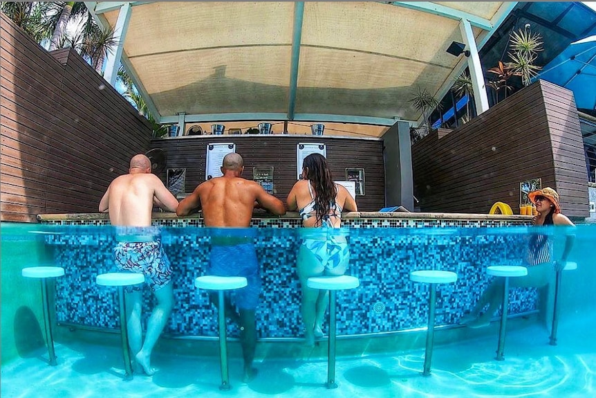 A semi-immersed image of four people in swimming costumes sitting on underwater barstools at a swim-up pool bar.