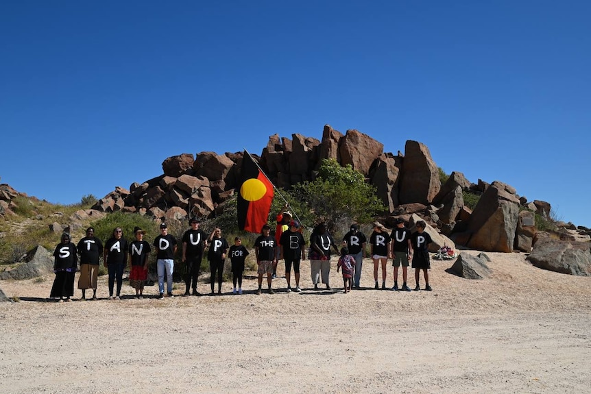 A group of people with letters on their shirts stand under an Aboriginal flag.