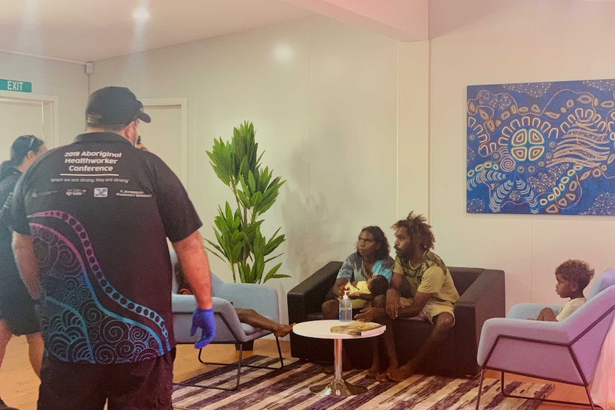 AN inside room with several Aboriginal people sitting on chairs, talking to health workers who have their back to the camera