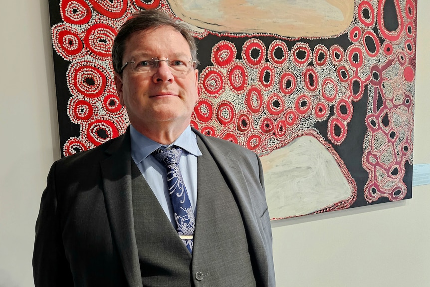 Bespectacled man wearing suit, tie and vest standing in front of Aboriginal dot painting