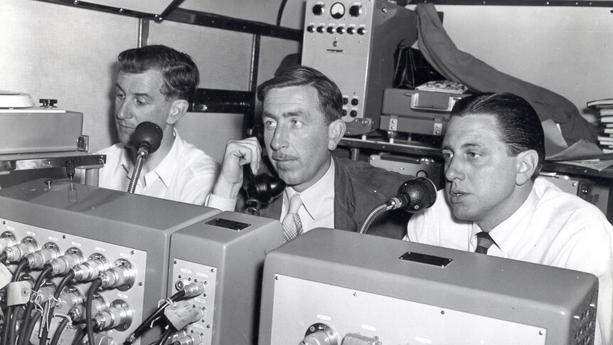 Three men sit in a cramped space surrounded by technical equipment.