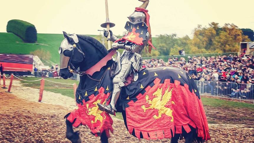 A knight dressed in medieval costume sits on a horse holding a lance while a crowd watches on behind.