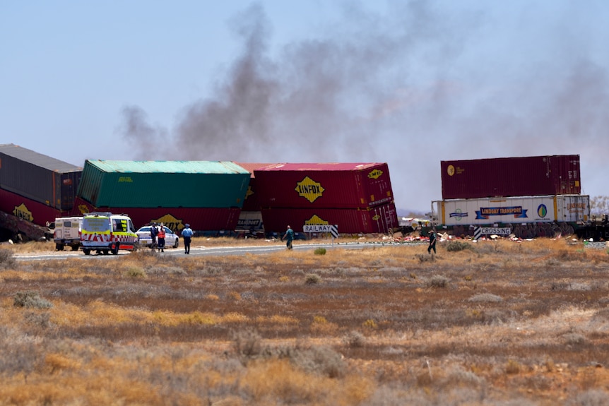 A tipped over freight train with grey smoke in the air and an ambulance and police car in front