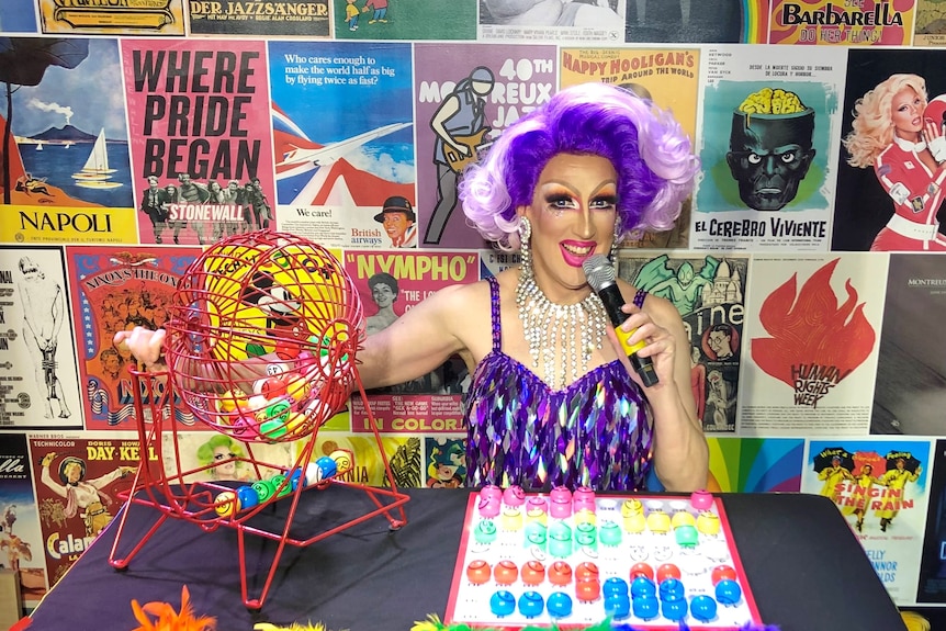 A brightly dressed drag queen sits at a table with bingo materials 