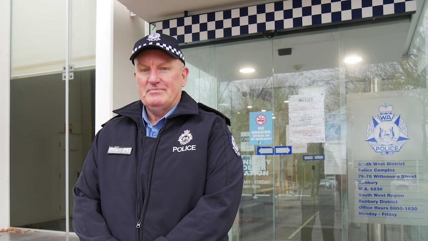 A police officer wearing a hat stands in front of glass doors at a police station