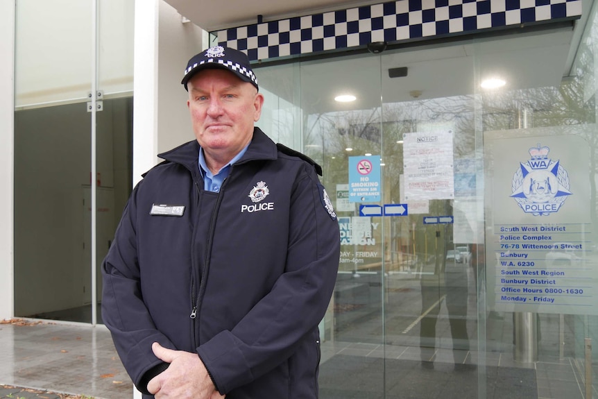A police officer wearing a hat stands in front of glass doors at a police station