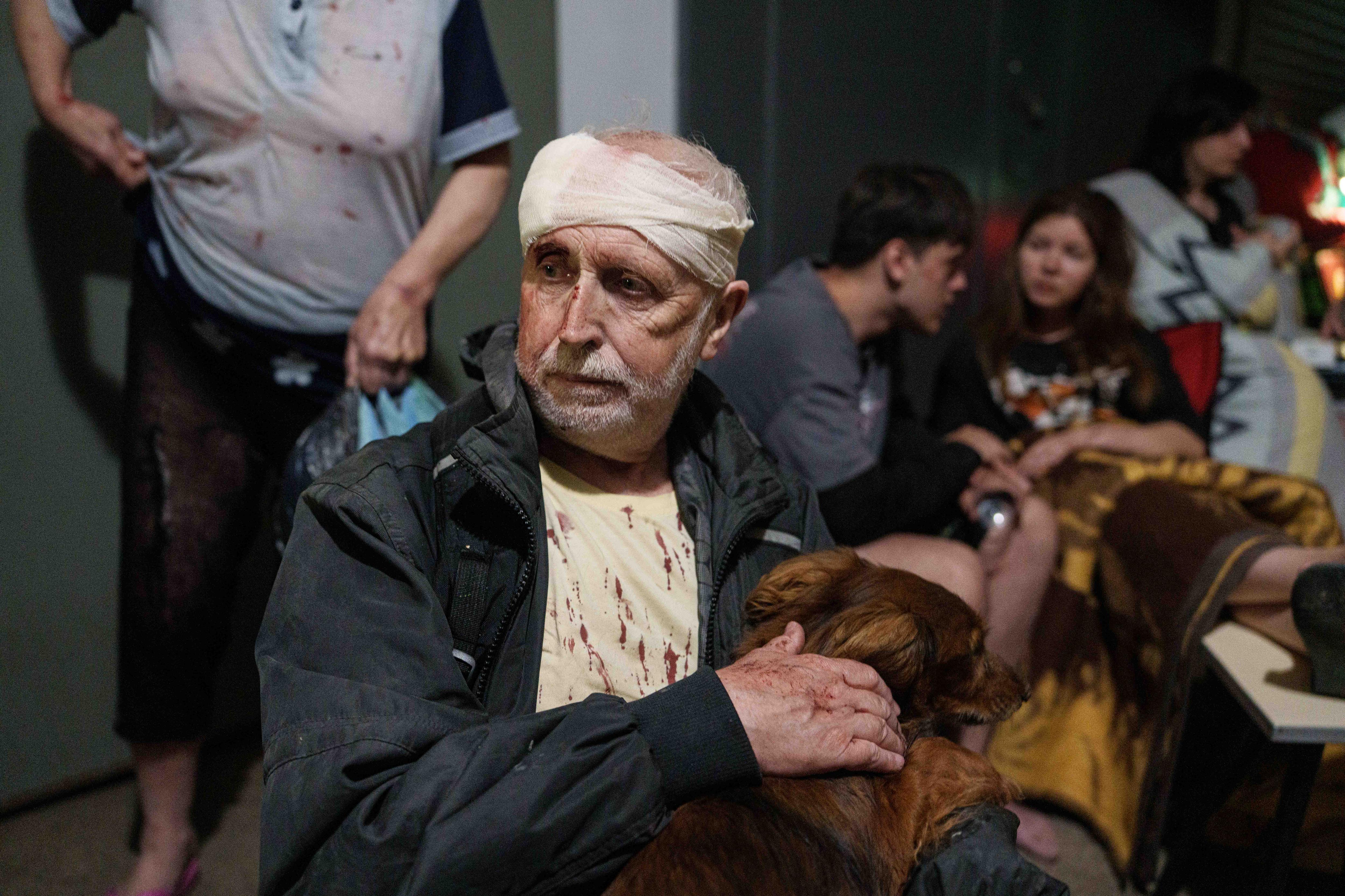 A man with a bandaged head with blood on his t-shirt pats his dog while sheltering in an underground bomb shelter.