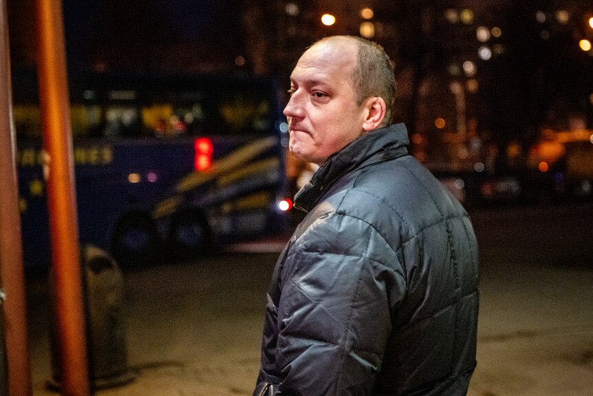 A man with tears in his eyes and a pained expression turns towards the camera. A bus is visible in the background