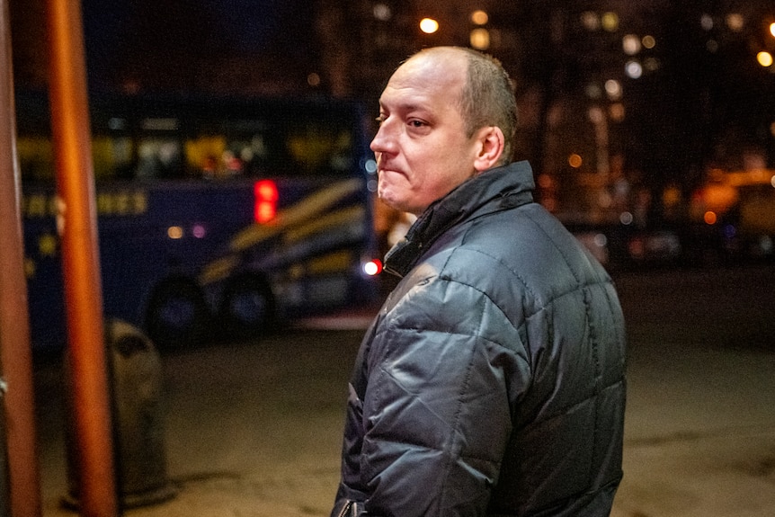 A man with tears in his eyes and a pained expression turns towards the camera. A bus is visible in the background