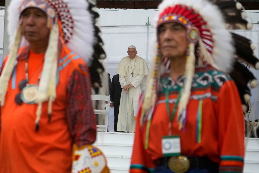 Two men in traditional dress are prictured out of focus and between them Pope Francis stands further away in robes. 