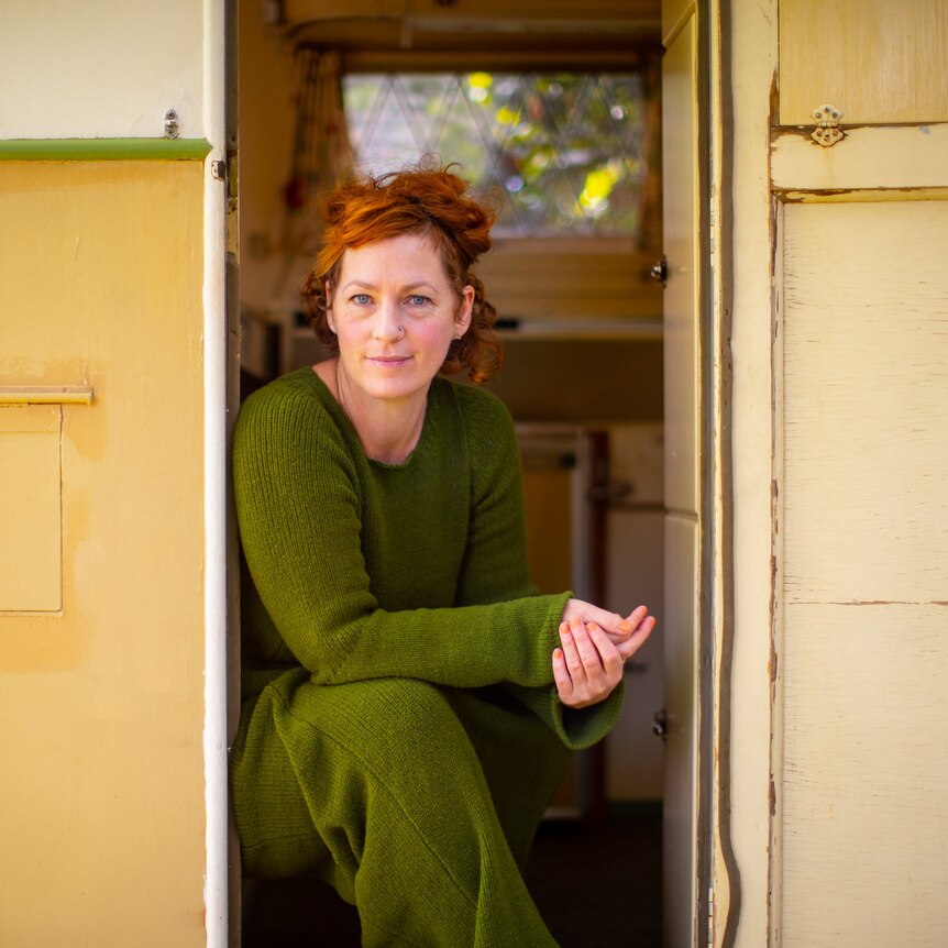 Jenny sits in the doorway of an old yellow caravan, wearing soft green knits and looking at the camera