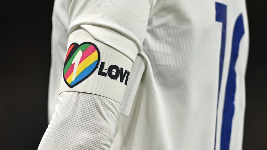 A white armband with a rainbow heart reading '1 Love' on a player's arm. 