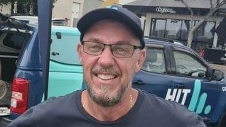 A bearded man standing in front of a branded vehicle, wearing a cap, spectacles and smiling.