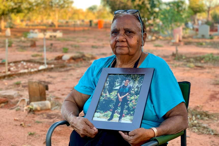 A woman in a blue shirt sits outside at a cemetery holding a framed photograph of a young man 