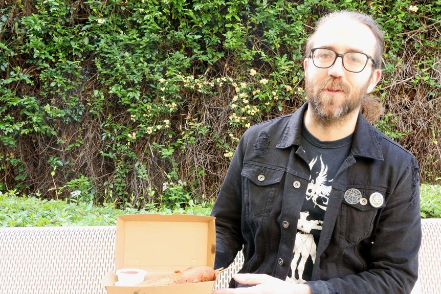 Photograph of a young man with beard and glasses holding a cardboard box with dagwood dogs and tomato sauce
