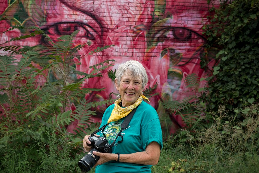 An older white female photographer with a camera around her neck, in front of a mural