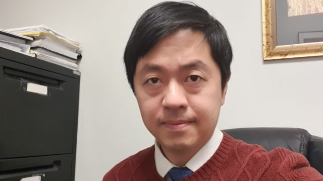 A selfie of a Hong Kong man with black hair in an office wearing a red knitted jumper. Cabinets and frames are in background.