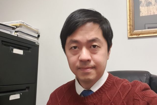 A selfie of a Hong Kong man with black hair in an office wearing a red knitted jumper. Cabinets and frames are in background.